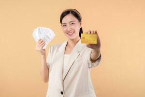 portrait of happy successful confident young asian business woman wearing white jacket holding cash money dollars and credit card standing over beige background. millionaire business, shopping concept photo