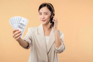 portrait of happy successful confident young asian business woman wearing white jacket holding smartphone and cash money dollar standing over beige background. millionaire business, shopping concept. photo