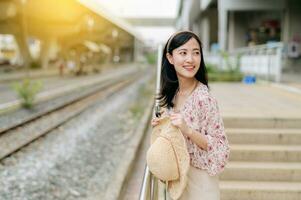 young asian woman traveler with weaving basket waiting for train in train station. Journey trip lifestyle, world travel explorer or Asia summer tourism concept. photo