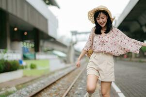 joven asiático mujer viajero con Costura cesta contento sonriente mirando a un cámara junto a tren ferrocarril. viaje viaje estilo de vida, mundo viaje explorador o Asia verano turismo concepto. foto