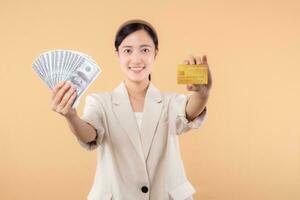 portrait of happy successful confident young asian business woman wearing white jacket holding cash money dollars and credit card standing over beige background. millionaire business, shopping concept photo