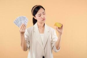 portrait of happy successful confident young asian business woman wearing white jacket holding cash money dollars and credit card standing over beige background. millionaire business, shopping concept photo