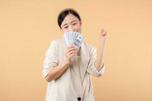 portrait of happy successful confident young asian business woman wearing white jacket holding cash money dollars standing over beige background. millionaire business, shopping concept. photo