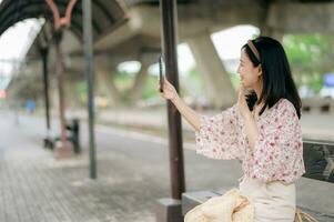 joven asiático mujer viajero con Costura cesta utilizando un móvil teléfono y esperando para tren en tren estación. viaje viaje estilo de vida, mundo viaje explorador o Asia verano turismo concepto. foto