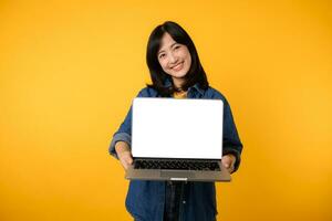 portrait young happy woman wearing yellow t-shirt and denim shirt holding laptop and point finger to screen isolated on yellow studio background. business technology application communication concept. photo