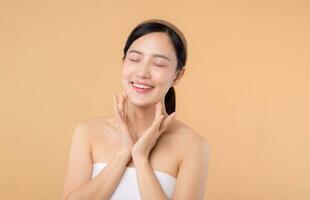 Beautiful girl asian model touching fresh glowing hydrated facial skin on beige background closeup. Beauty face young woman with natural makeup and healthy skin portrait. Skin care concept photo