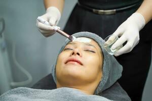 Closeup cosmetologist is applying facial white mask cream on woman client face in beauty clinic. photo