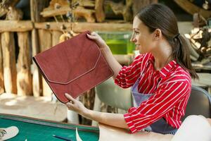 joven hembra cuero bienes fabricante mirando y cheque el pulcritud de el cueros bolso antes de vender a cliente en taller. foto