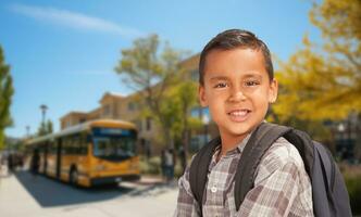 Cute Young Hispanic Boy Wearing a Backpack Near a School Bus on Campus. photo
