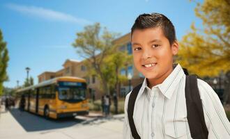 Happy Young Hispanic Boy Wearing a Backpack Near a School Bus on Campus. photo