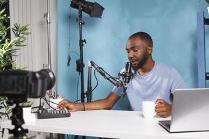 African american blogger creating professional video content with camera and volume controller. Vlogger using sound mixer console while live streaming and drinking coffee in studio photo