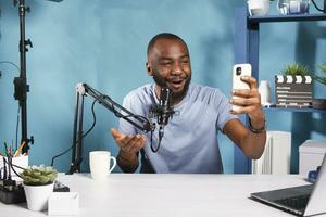 Blogger creating engaging content with smartphone and speaking to subscribers. Smiling african american vlogger talking while live streaming on social media network with mobile phone photo