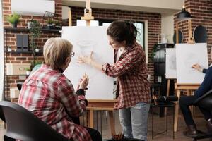 Female art teacher standing near easel explaining senior woman how to draw still life objects with pencil, teaching people drawing techniques during group art class. Learning new things in retirement photo