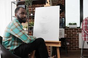 africano americano chico sentado a caballete en salón de clases y mirando a cámara, estudiando multa Arte. personas participativo en dibujo clase maestra, aprendizaje varios tecnicas centrado alrededor lápiz dibujando foto