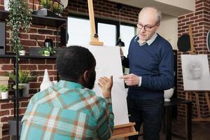 People connecting while learning new skills, two diverse men looking at easel with canvas discussing artwork while studying pencil drawing together at group art class. Sketching lesson photo