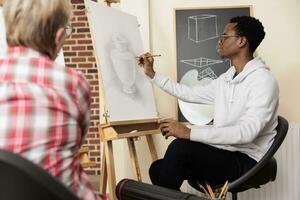 African American millennial guy drawing flower vase on canvas with pencil, learning to draw in group, spending time focusing on and creating art. People improving creativity with creative hobby photo