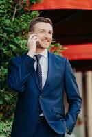 Upbeat businessman in blue suit making a call with a smile, standing by greenery and red awning photo