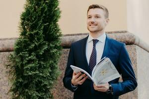 Smiling businessman in a blue suit reading a magazine outdoors with greenery in the backdrop. photo