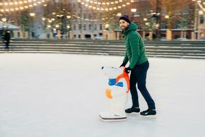 Joyful man skating with a bear-shaped aid on a festive ice rink in the evening photo