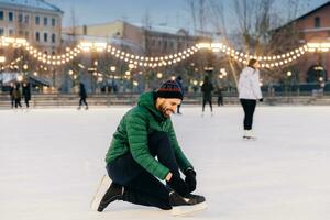 Man tying his ice skates on a festive lit rink at dusk, with skaters behind. photo