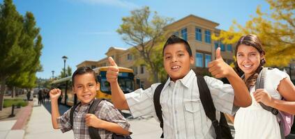 Excited Young Hispanic Boy Wearing a Backpack Giving Two Thumbs Up Surrounded by His Friends on Campus photo