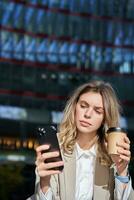 Vertical shot of young corporate woman drinks her coffee and looks at mobile phone, texts message, stands outside in city photo