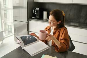 retrato de joven mujer disfrutando su fin de semana, leyendo un libro y Bebiendo té a hogar, vistiendo lentes y casual ropa foto