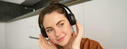 Close up shot of cute young woman in wireless headphones, listens to music and smiles, relaxes at home with favourite songs playing on her playlist photo