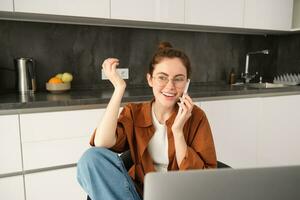 Excited young woman talking on mobile phone in front of laptop, sitting in kitchen with happy face expression, having a conversation photo