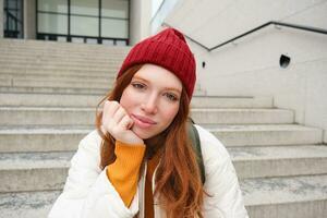 Close up portrait of beautiful redhead girl in red hat, urban woman with freckles and ginger hair, sits on stairs on street, smiles and looks gorgeous photo