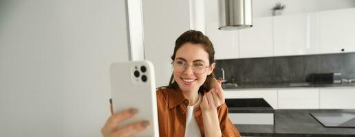 Cute young woman taking selfie in her flat, showing heart sign, using smartphone camera making lovely pictures for social media photo