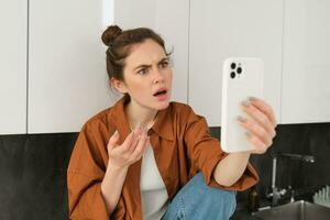 Portrait of woman with confused face, talking at smartphone, video chats, expresses frustration and disappointment photo