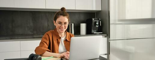 Portrait of young woman working from home, sets up workplace in kitchen, using laptop. Student doing homework on computer photo