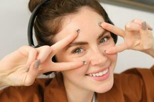 Close up portrait of beauty girl, listens music in wireless headphones, shows peace signs near face and smiles at camera photo