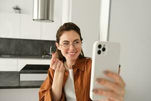 Cute young woman taking selfie in her flat, showing heart sign, using smartphone camera making lovely pictures for social media photo