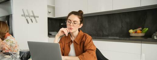 retrato de joven mujer, estudiante estudiando remoto, conecta a en línea lección en computadora portátil, sentado a hogar y trabajo independiente, trabajando desde su cocina foto