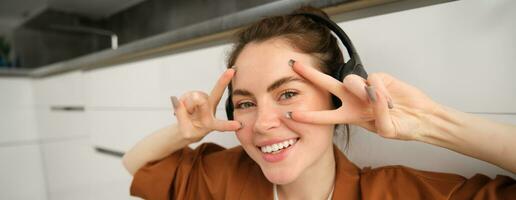 Close up of carefree young woman, laughing and smiling, showing peace, vsign gesture, listening to music in wireless headphones photo