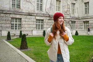 Young smiling redhead woman walking in beautiful city attractions, wearing backpack, red hat and coat, looking around with happy face photo