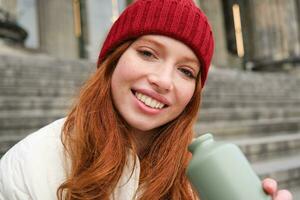 Young redhead female tourist rests during her trip, opens thermos and drinks hot tea, having a break after sightseeing photo