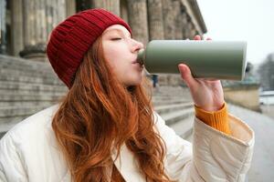 Young redhead female tourist rests during her trip, opens thermos and drinks hot tea, having a break after sightseeing photo