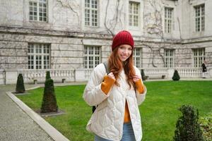 retrato de sonriente chica, mochilero en Saco y rojo sombrero, vistiendo calentar ropa para turista viaje alrededor Europa en invierno, caminando alrededor histórico edificio foto