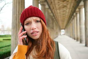 Portrait of redhead girl in red hat, calls someone, listens to voice message with concerned, confused face expression, using smartphone photo