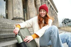 Young redhead female tourist rests during her trip, opens thermos and drinks hot tea, having a break after sightseeing photo