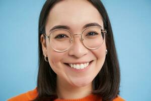 cerca arriba retrato de hermosa joven asiático mujer en anteojos, sonriente y mirando feliz, molesto nuevo gafas a ópticos almacenar, soportes terminado azul antecedentes foto