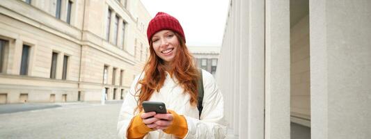Beautiful smiling girl, tourist with backpack, holding smartphone, using map on mobile phone application, looking for sightseeing in internet app, standing outdoors photo