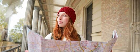 sonriente joven pelirrojo mujer en rojo sombrero, mira a papel mapa a Mira para turista atracción. turismo y personas concepto. niña explora ciudad, intentó a encontrar camino foto