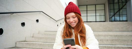 Stylish european girl with red hair, sits on public stairs with smartphone, places online order, sends message on mobile phone social app, smiles happily photo