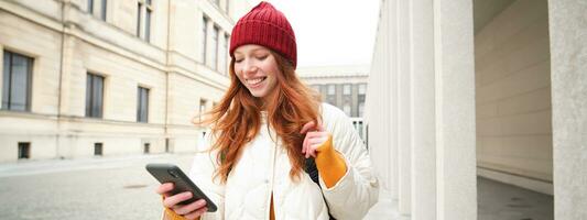 Beautiful smiling girl, tourist with backpack, holding smartphone, using map on mobile phone application, looking for sightseeing in internet app, standing outdoors photo