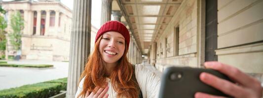 Cute young redhead woman takes selfie on street with mobile phone, makes a photo of herself with smartphone app on street