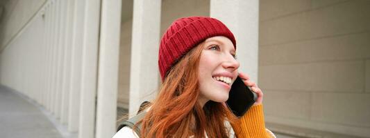 contento pelirrojo mujer, niña con teléfono negociaciones, tiene conversacion en móvil aplicación, usos Internet a llamada extranjero con teléfono inteligente aplicación, riendo y sonriente foto